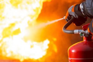 istockphoto-1327086620-612×612 Close-up showing a person pointing the nozzle of a fire extinguisher at the base of a large fire.
